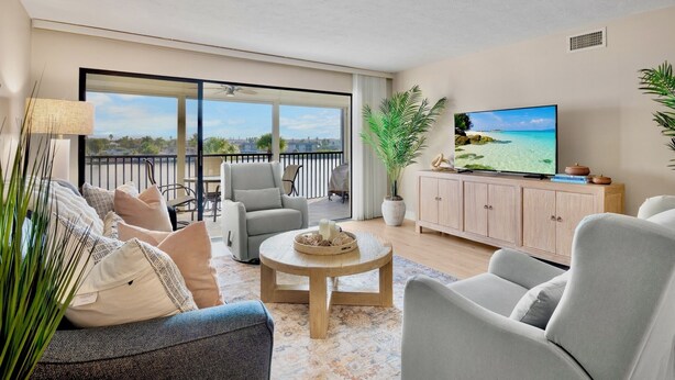 A modern living room with a large TV, comfortable seating, a balcony with a view, and a potted plant. - Midnight Cove II – Unit 133 (Siesta Key)