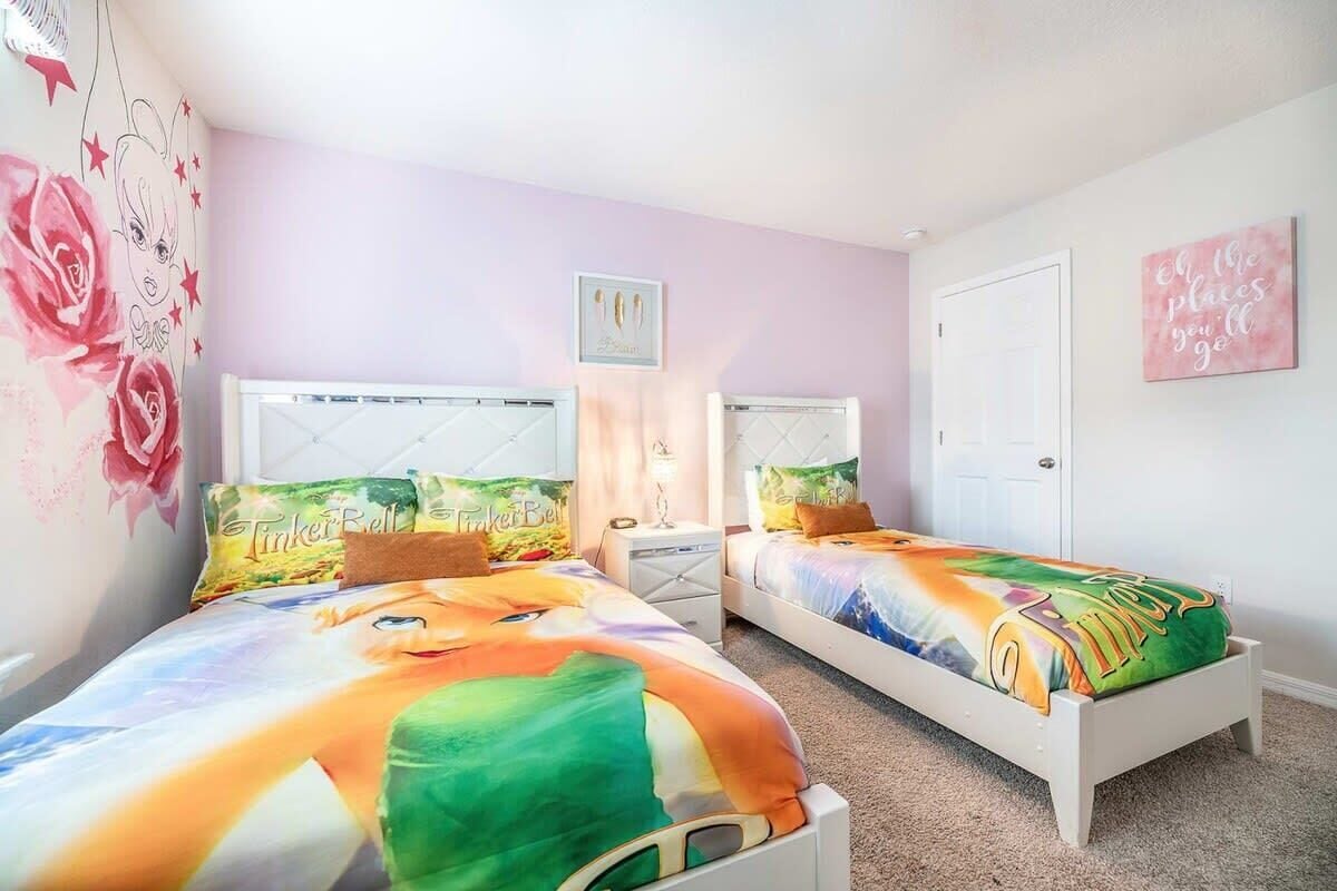 A bedroom with two beds, each with a colorful bedspread featuring a cartoon character. There is a nightstand with a lamp between the beds, and a door to the right.