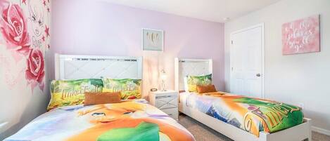 A bedroom with two beds, each with a colorful bedspread featuring a cartoon character. There is a nightstand with a lamp between the beds, and a door to the right.