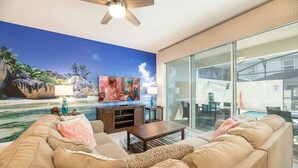 A living room with a large TV, a ceiling fan, and a glass door leading to a pool area. - Sunny Retreat-colorful Home, Pool, Great Location (Kissimmee)