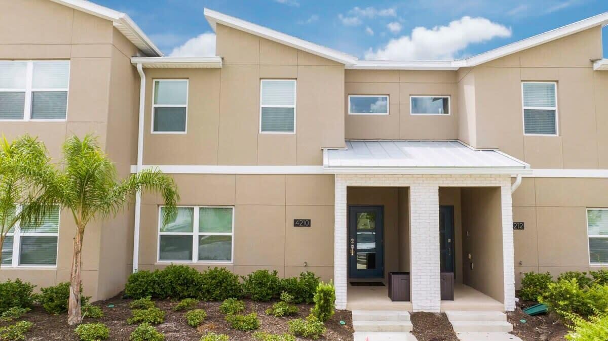 A multi-story residential building with a beige exterior, numbered 4210 and 212, featuring a central entrance with a covered porch and a small garden area with shrubs and a palm tree.