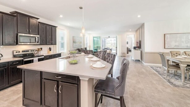 A modern kitchen with dark cabinetry, a central island, and a dining area with a long table and chairs. - Stunning Retreat, Private Pool, Perfect Location (Kissimmee)