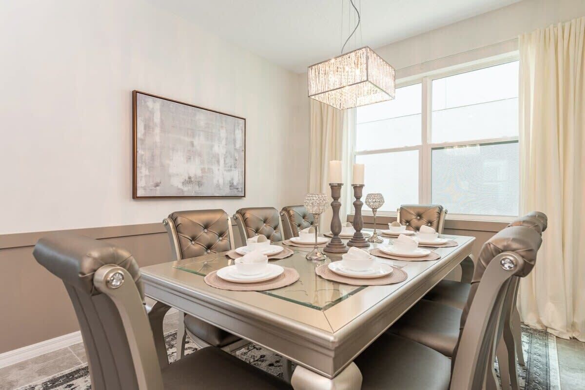 A dining room with a glass-top table, cushioned chairs, a chandelier, and a large window with curtains.