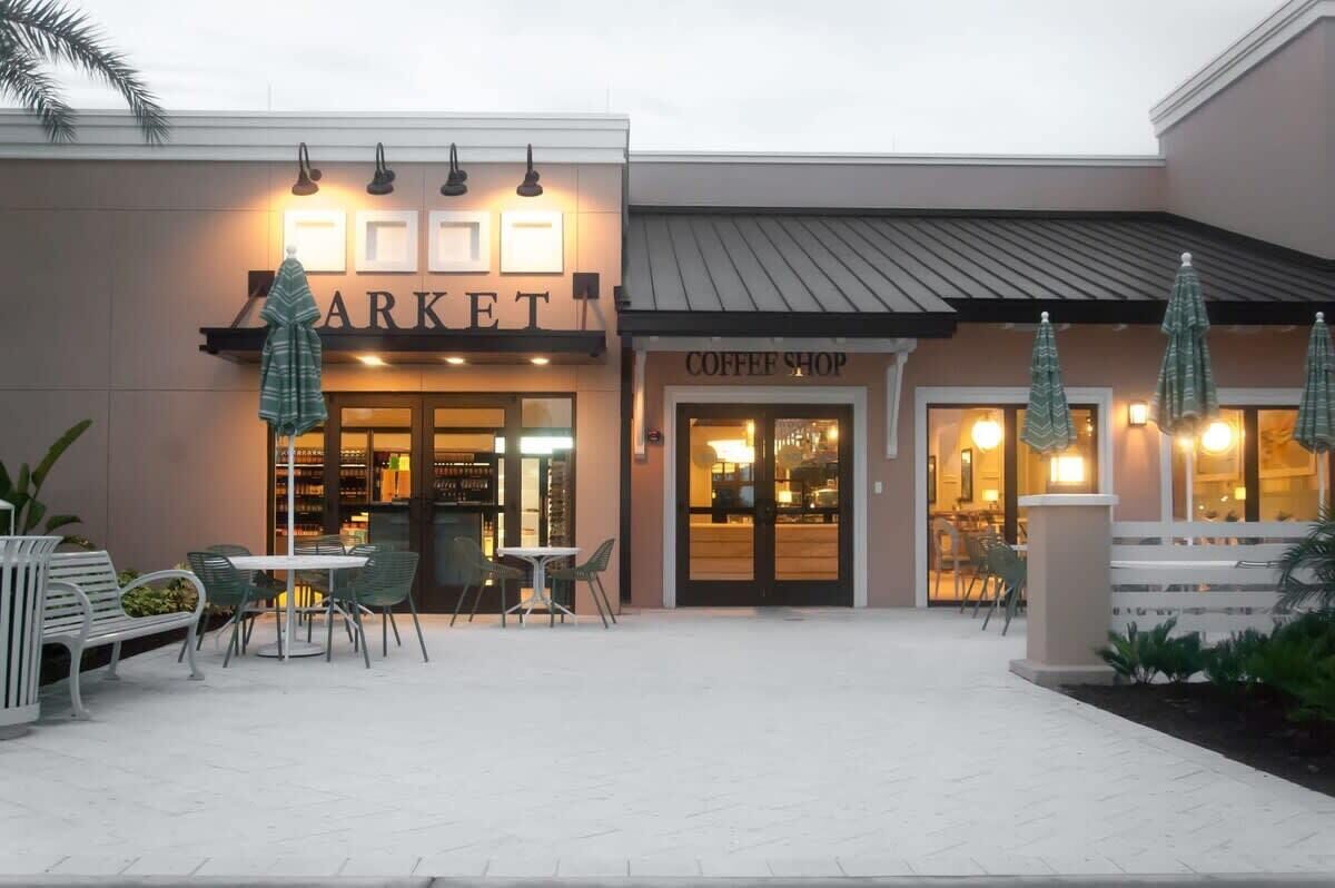 Un edificio con un letrero que dice 'Market' y otro que dice 'Coffee Shop'. Hay mesas y sillas al aire libre, y sombrillas.