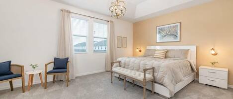 Un dormitorio con cama, mesitas de noche, un banco, una silla, una pequeña mesa y una ventana con cortinas.