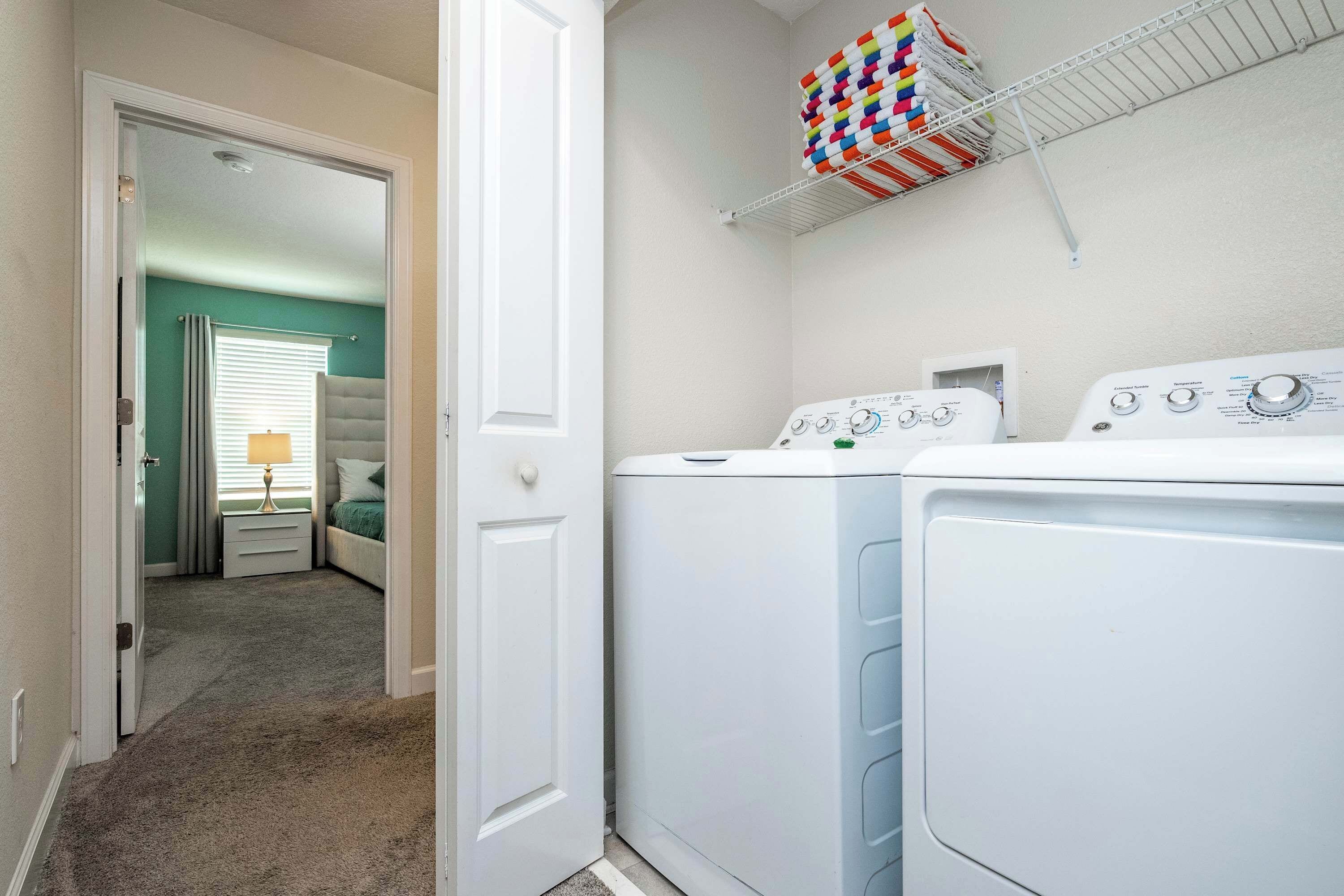 A laundry area with two white washing machines, a shelf with colorful storage bins, and a door leading to a bedroom with a bed and a nightstand.