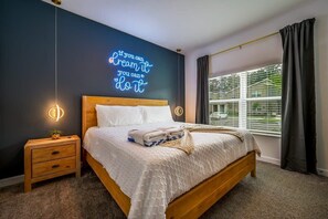 A neatly made bed with a headboard, a bedside table with a lamp, a wall with a neon sign, and a window with blinds. - Ultimate Luxury Home-private Pool, Themed Bedroom (Kissimmee)