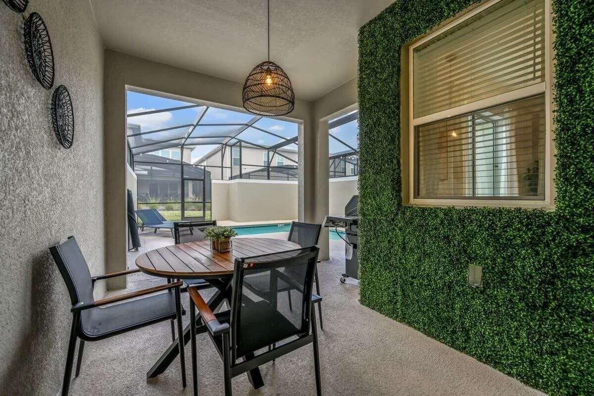 Eine Terrasse mit rundem Tisch und Stühlen, einer hängenden Leuchte, einem Fenster mit Jalousien und einer mit Grünpflanzen bewachsenen Wand.
