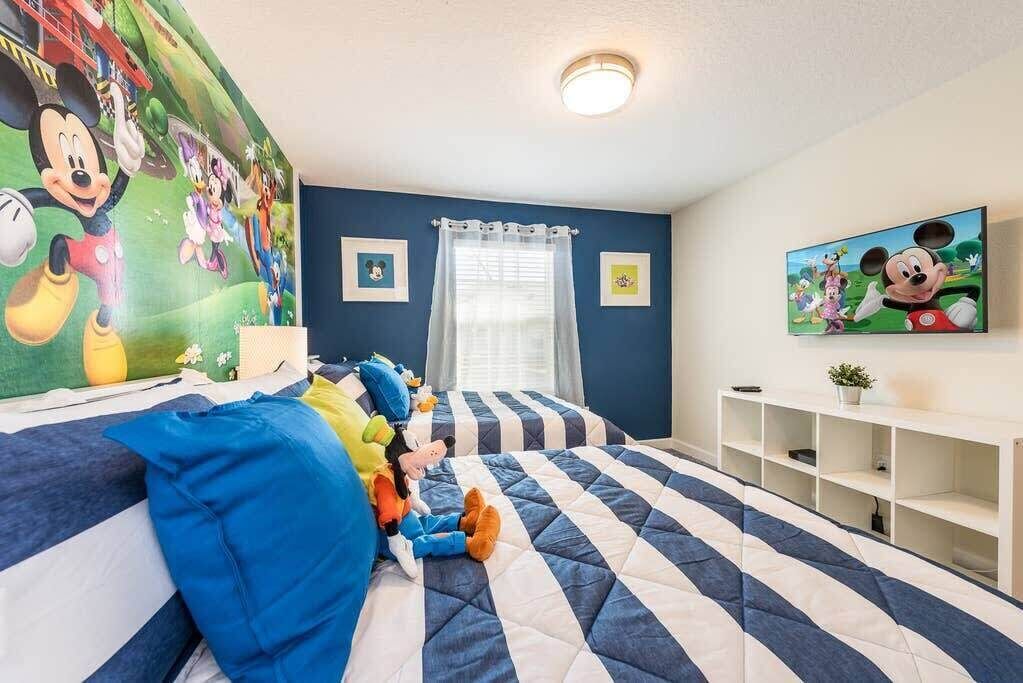 Une chambre avec une fresque représentant Mickey Mouse, un lit à rayures, une console blanche et une fenêtre avec des rideaux.
