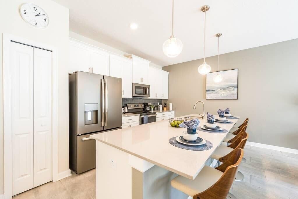 Una cocina moderna con armarios blancos, electrodomésticos de acero inoxidable, una isla central con asientos, iluminación colgante y un reloj en la pared.