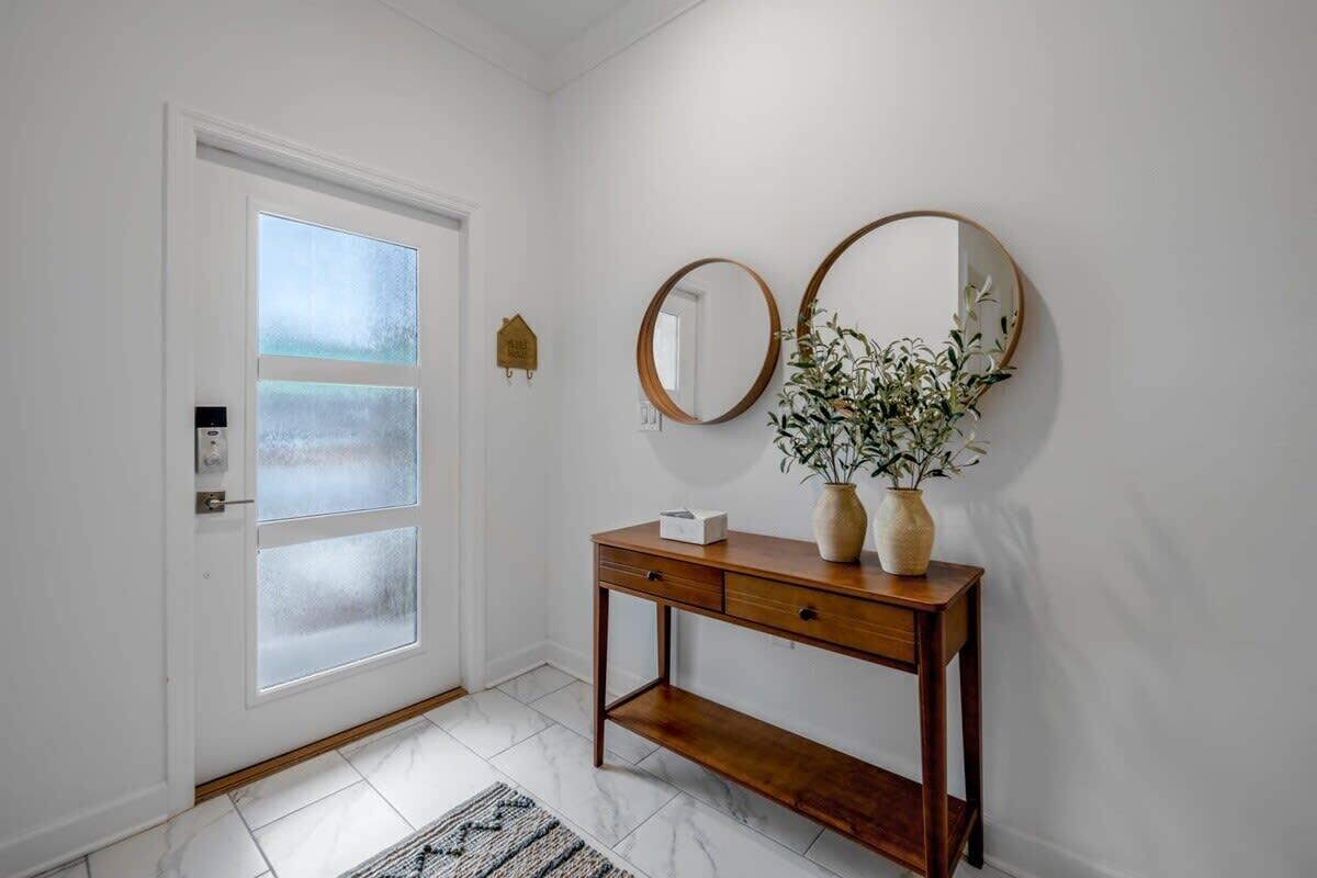 Un pasillo con una puerta blanca, una consola de madera con un florero y un espejo, y una pequeña decoración con forma de casa en la pared.