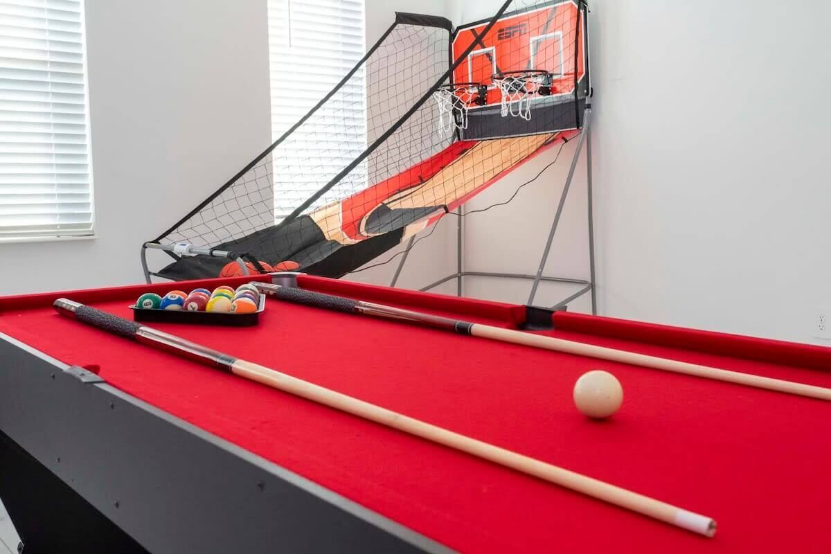 A pool table with balls and cues, a window with blinds, and a basketball hoop.