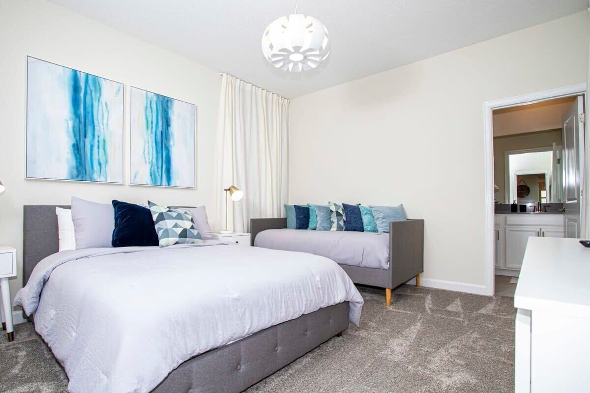 A modern bedroom with two beds, a large window with curtains, and a kitchen visible through an open door.