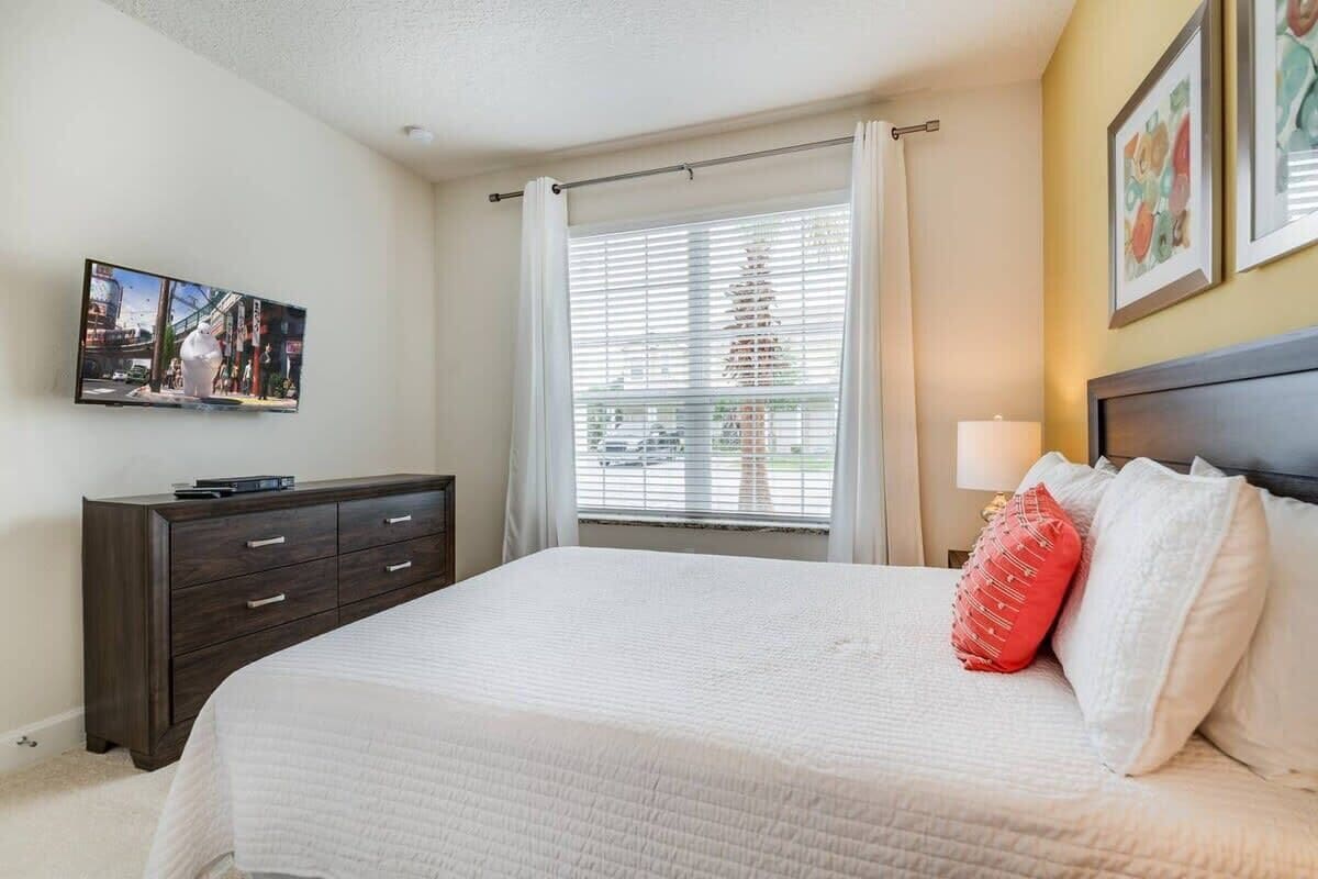 A bedroom with a bed, a dresser, a window with blinds, and a wall-mounted TV.