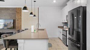 Cocina moderna con una isla grande, electrodomésticos de acero inoxidable, iluminación colgante y un televisor de pantalla plana.