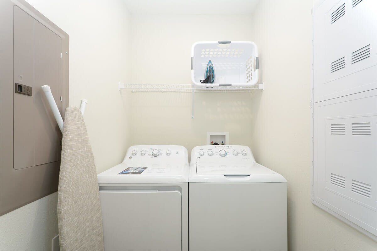 Un espace buanderie comprenant deux machines à laver blanches, un étendoir et un meuble de rangement avec un fer à repasser.
