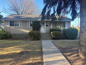 A single-story house with a front porch, a mailbox, and a small front yard with a lawn and two bushes. - Monthly Rentals (Edmonton)