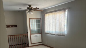 A room with a ceiling fan, a window with patterned curtains, a wooden railing, and a built-in cabinet. - Monthly Rentals (Edmonton)