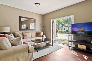 A living room with a beige sofa, a coffee table, a TV on a stand, and a large window with a view of a building and trees. - Welcoming 4 BR Sleeps 10 Guests (Kissimmee)
