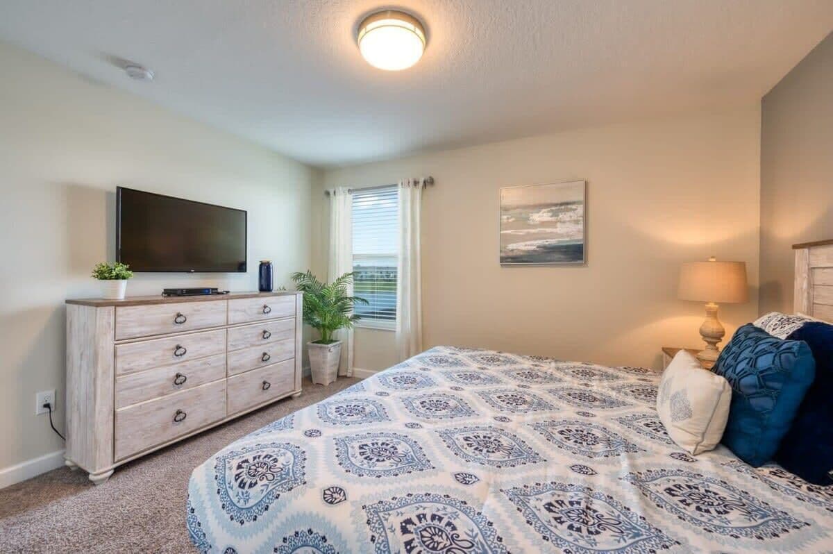 Une chambre à coucher avec un lit, une commode, un téléviseur, une fenêtre avec des rideaux et un tableau au mur.
