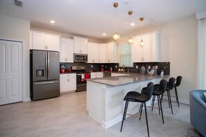 A modern kitchen with white cabinetry, a stainless steel refrigerator, a central island with bar stools, and pendant lighting. - Ultimate Themed Luxury - Game Room, Theater, Pool (Kissimmee)