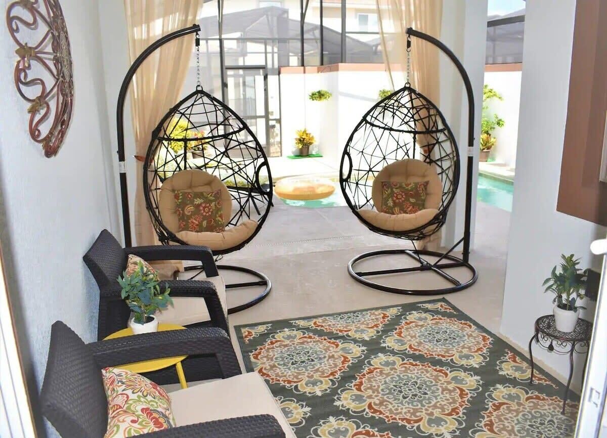 A room with two hanging chairs, a sofa, a patterned rug, and a view of a pool.