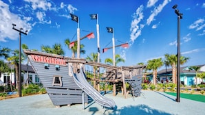 A playground with a slide and climbing structures, surrounded by palm trees and a clear blue sky. - Stylish Family Retreat With Themed Bedrooms (Kissimmee)