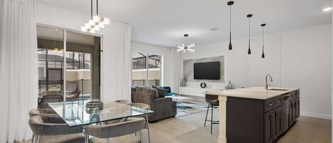 Una cocina moderna con una isla central, un comedor con mesa de vidrio y una sala de estar con sofá y televisor.