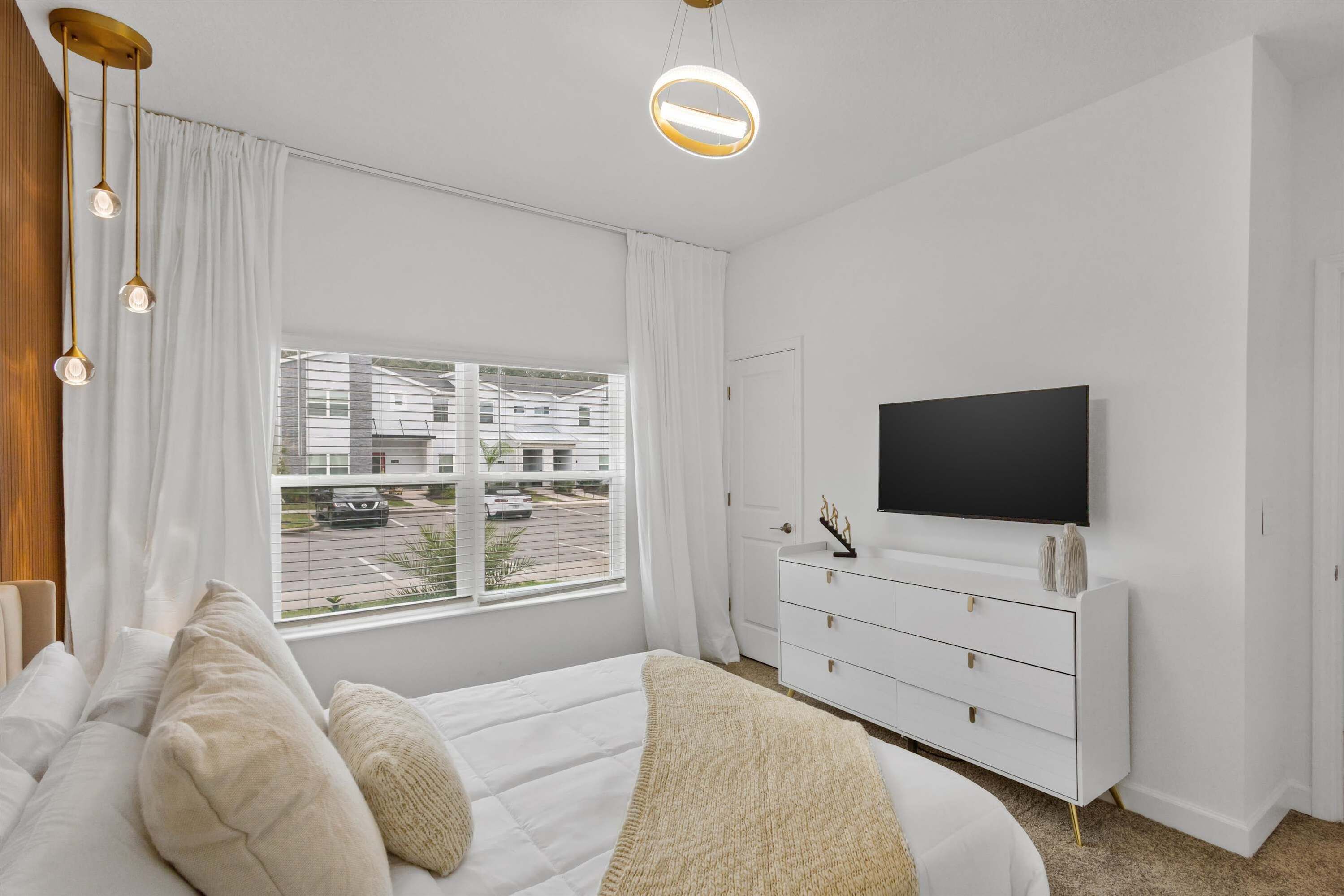 A bedroom with a bed, a dresser, a TV, and a window with curtains.