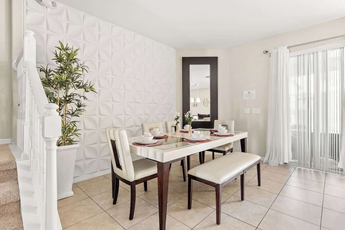 Uma área de refeições com uma mesa branca posta para quatro pessoas, cercada por cadeiras. Um vaso com planta está à esquerda, e um banco à direita.