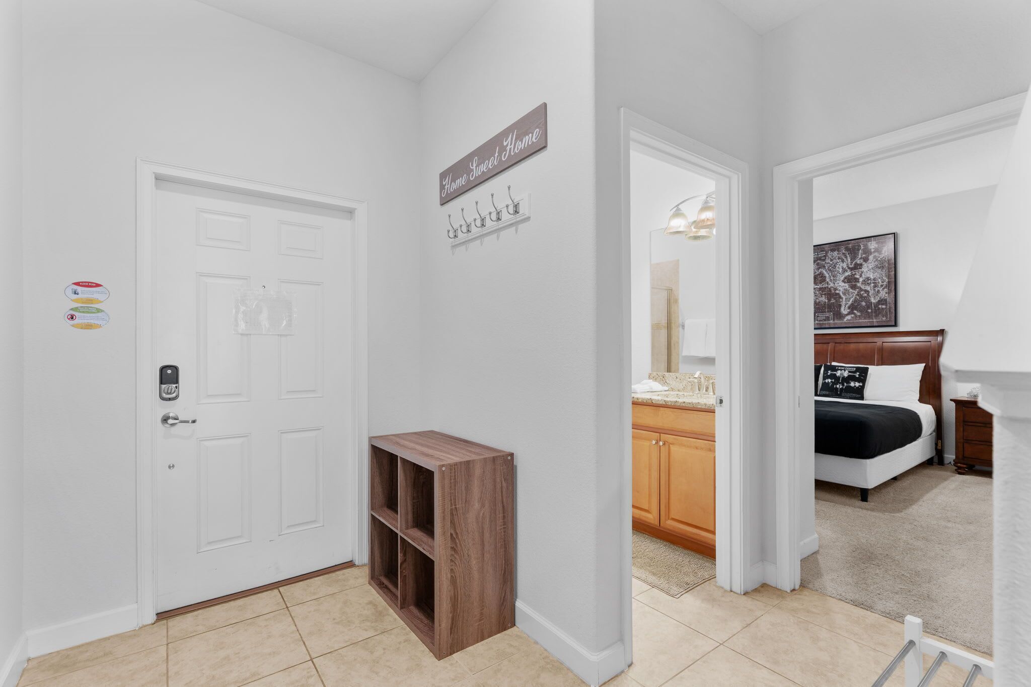 A hallway with a white door, a small wooden shelf, a sign reading 'Home Sweet Home,' and a bedroom with a bed and a nightstand.