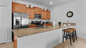 A kitchen with a stainless steel refrigerator, a microwave, wooden cabinets, a granite countertop, a sink, and two bar stools. - Townhome 4BR With Pool at Storey Lake (Kissimmee)