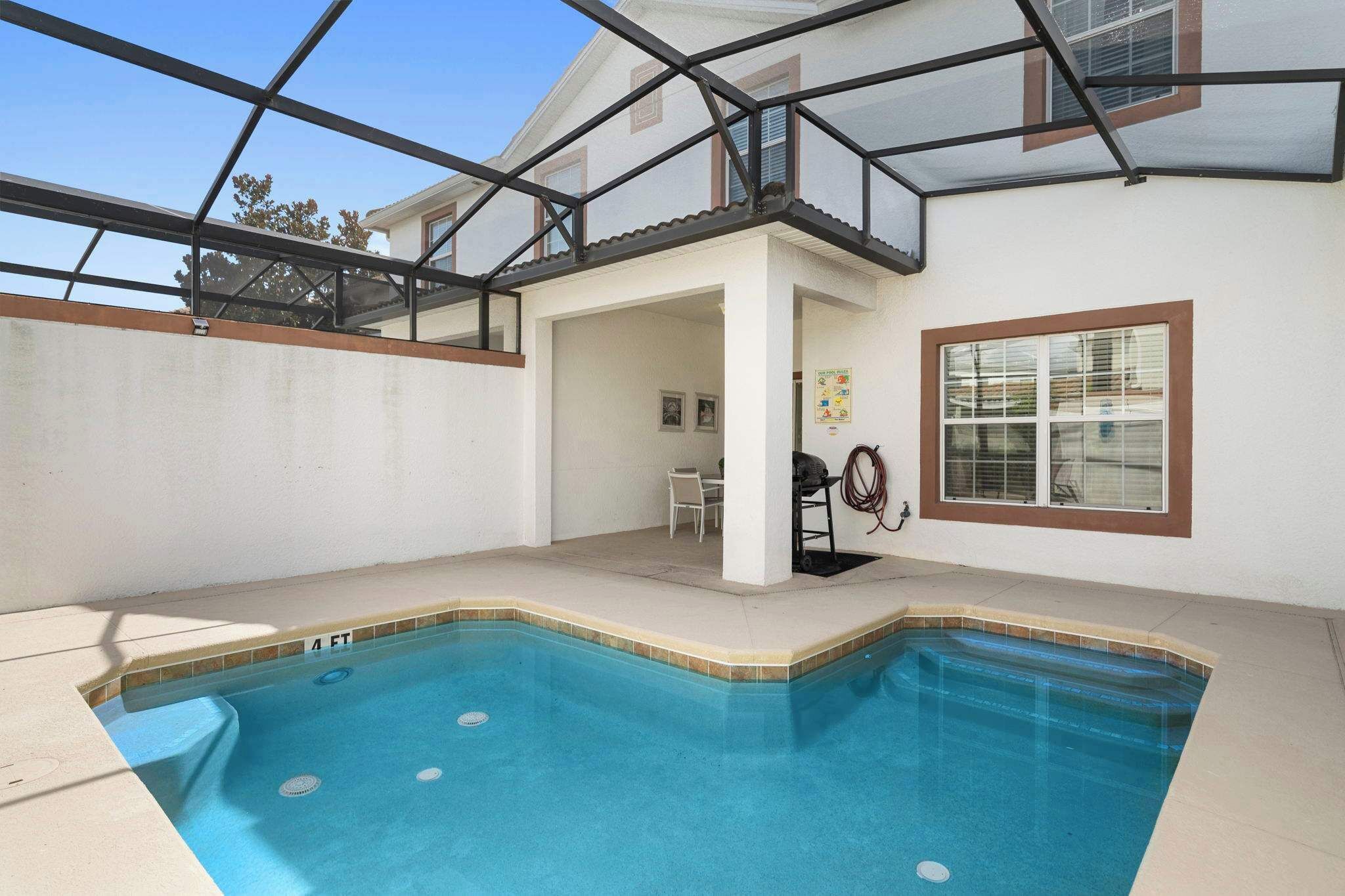A swimming pool with a glass enclosure, a patio area with a table and chairs, and a window with blinds.