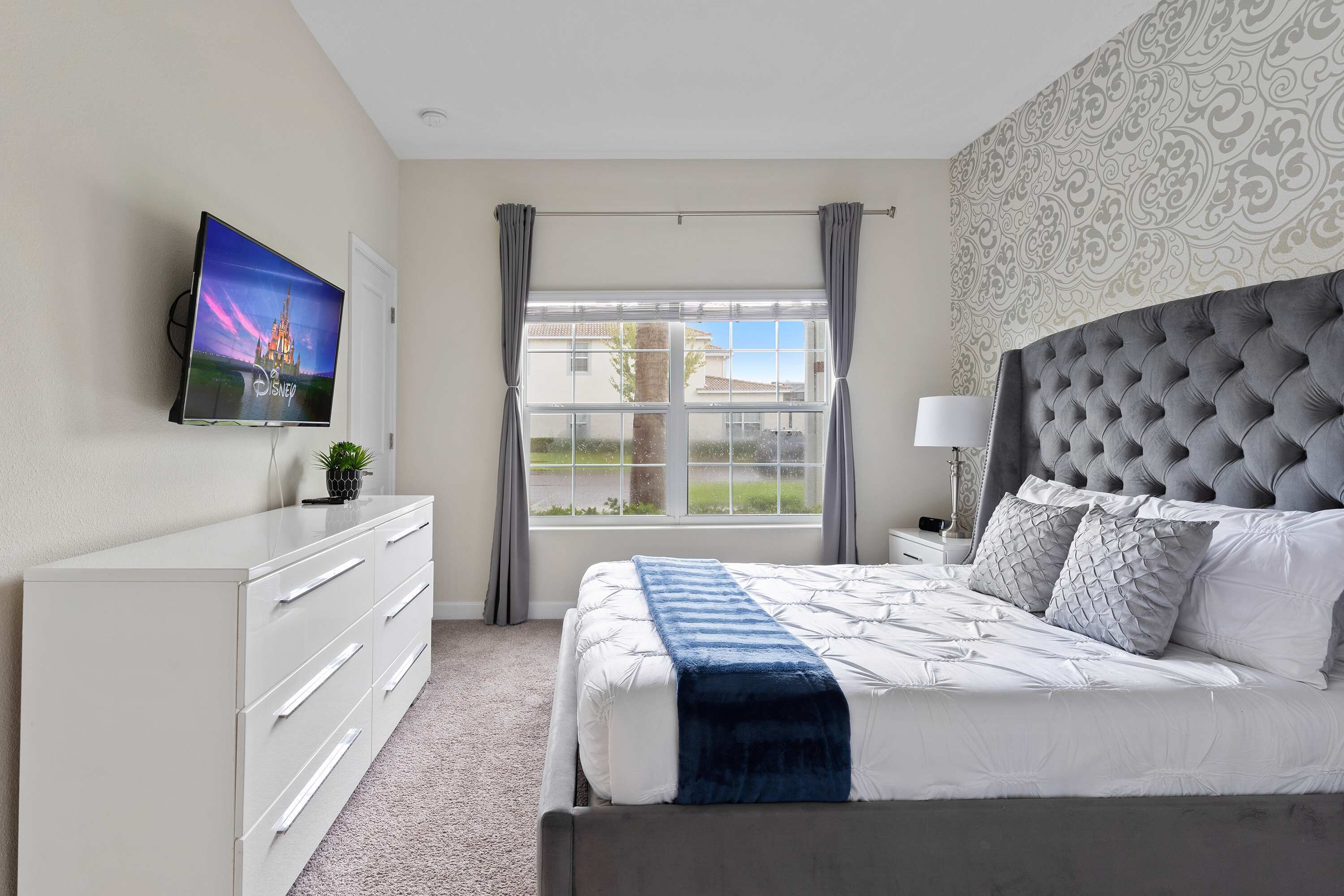 A bedroom with a bed, a TV on the wall, a dresser, and a window with curtains.