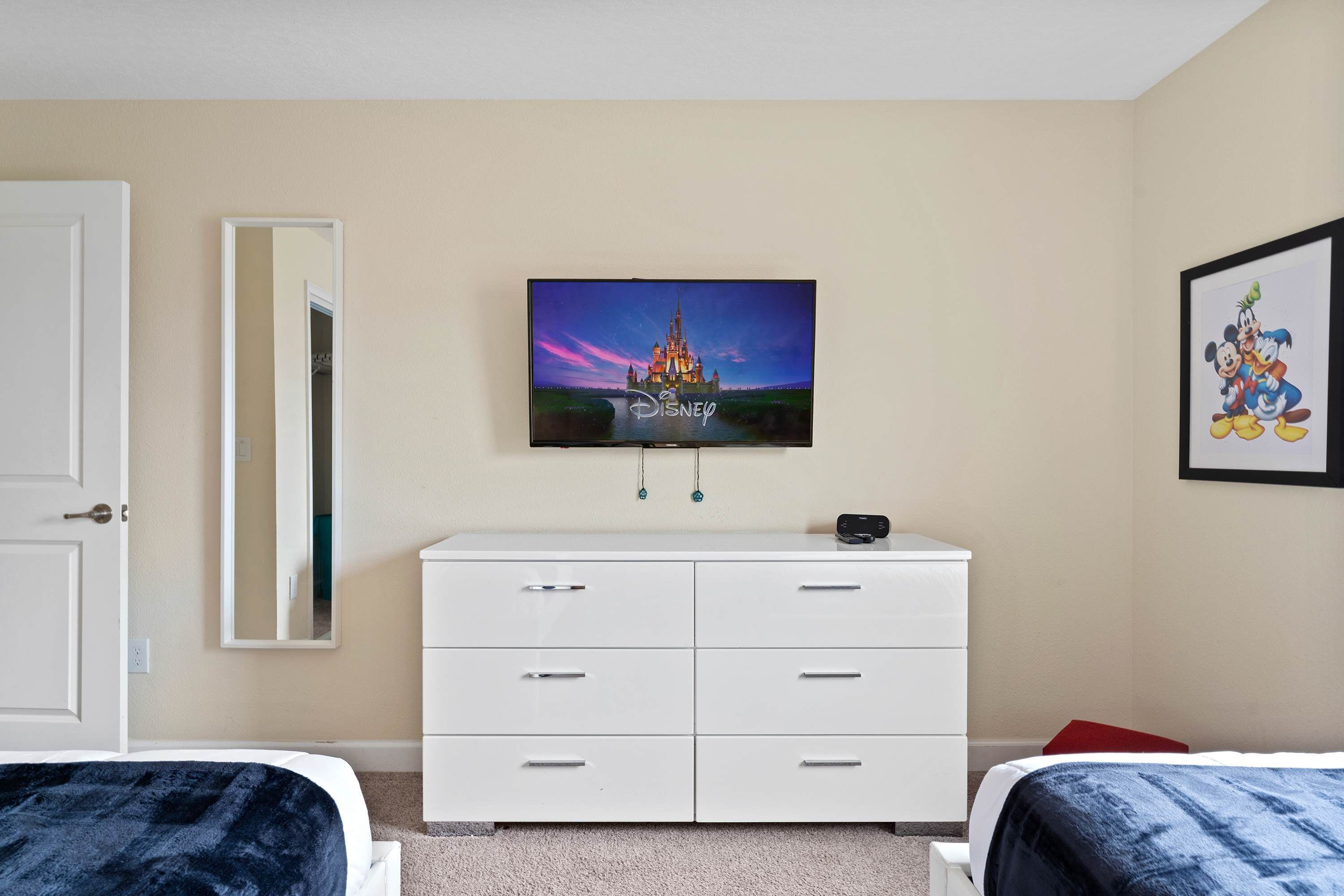 Een slaapkamer met een televisie waarop een Disney-kasteel te zien is, een witte dressoir, twee bedden, een spiegel en een ingelijst foto aan de muur.