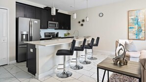 Una cocina moderna con armarios oscuros, electrodomésticos de acero inoxidable, una isla blanca con taburetes de bar, luces colgantes y una escultura decorativa sobre una mesa.