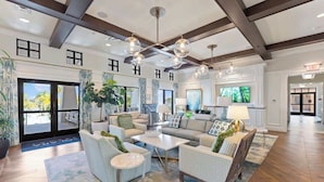 A spacious lobby with a high ceiling, a central seating area with sofas and chairs, a coffee table, and a large TV screen. There are potted plants and a blue rug on the floor. - Stunning 7BR Home With Themed Bedrooms (Kissimmee)