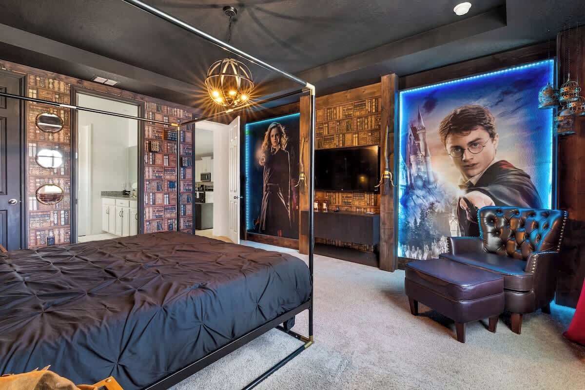 A bedroom with a large bed, a seating area with a chair and ottoman, a bookshelf, and a TV.
