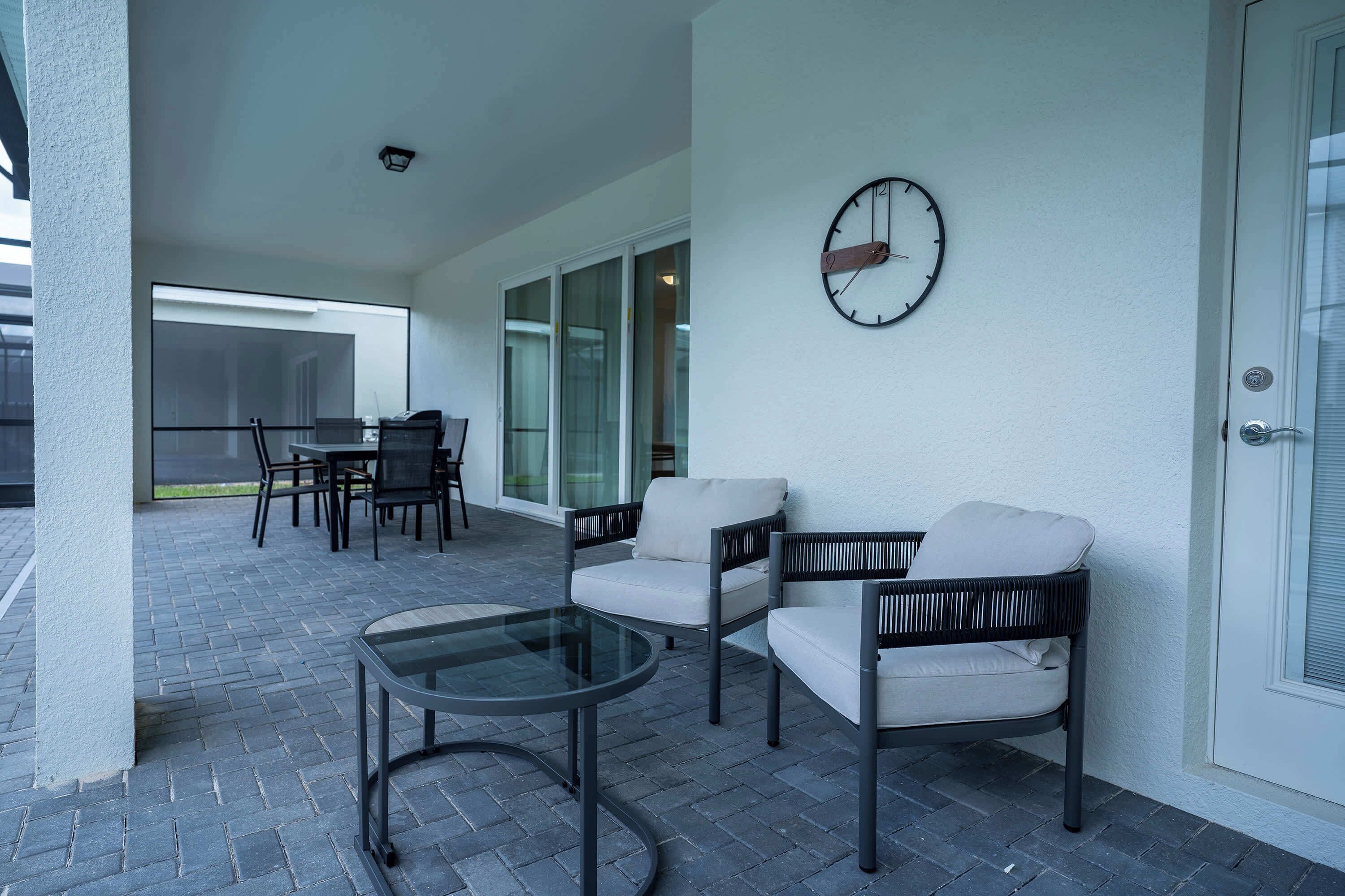 A covered patio with a glass table, two chairs, and a clock on the wall.