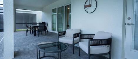 A covered patio with a glass table, two chairs, and a clock on the wall.