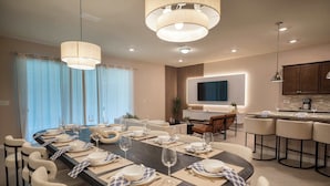 Uma área de refeições moderna com uma mesa comprida posta para seis pessoas, rodeada de cadeiras, e uma cozinha com banquetas.
