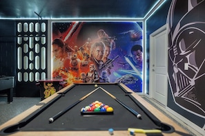 Una mesa de billar con una disposición colorida de bolas, una vara de billar y un fondo con un mural con temática espacial que presenta personajes y naves espaciales.
