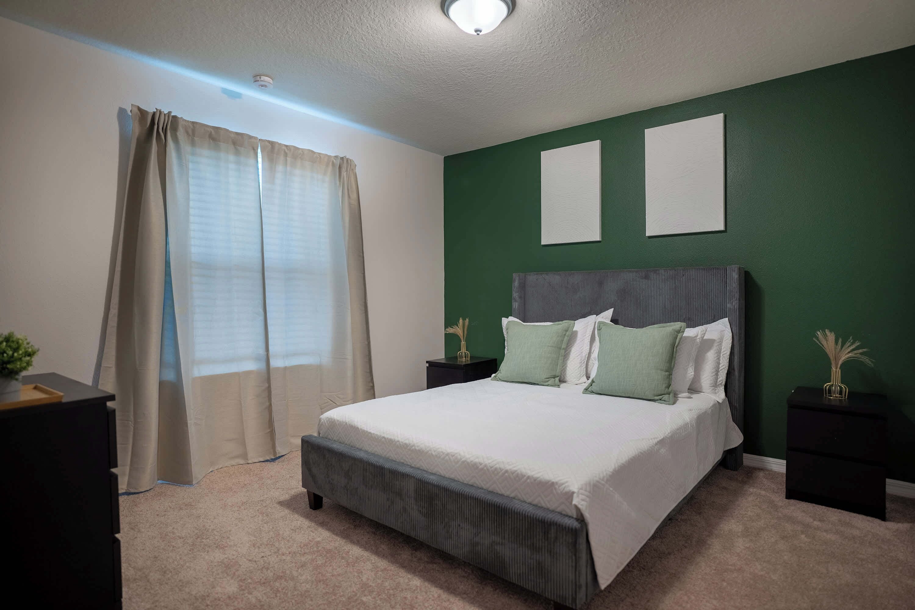 Une chambre à coucher avec un lit, deux tables de chevet, une commode, une fenêtre avec des rideaux et un mur vert.