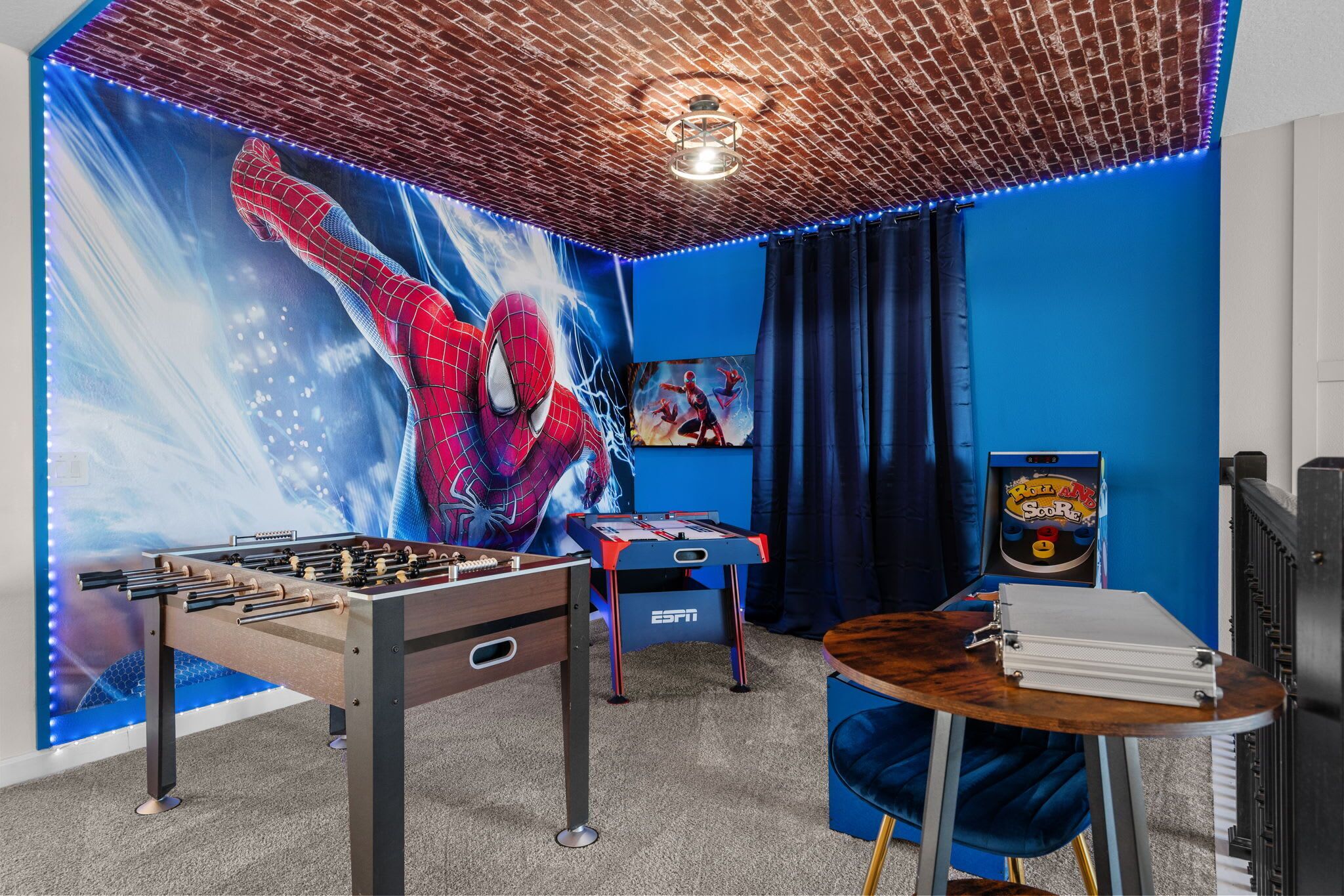 Una habitación con una mesa de fútbol de mesa, una mesa con un juego de mesa, una pared azul con un mural de Spider-Man y un techo con diseño de ladrillo.