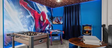 Una habitación con una mesa de fútbol de mesa, una mesa con un juego de mesa, una pared azul con un mural de Spider-Man y un techo con diseño de ladrillo.