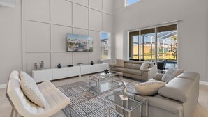A spacious living room with a large window, a sofa, a coffee table, and a TV mounted on the wall. - Stylish 5BR Home With Themed Bedrooms (Kissimmee)