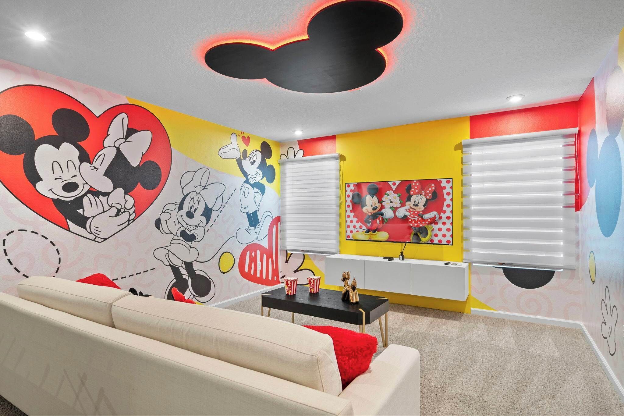 A room with a large Mickey Mouse mural, a white sofa, a red throw pillow, and a black coffee table with two red and white striped cups.