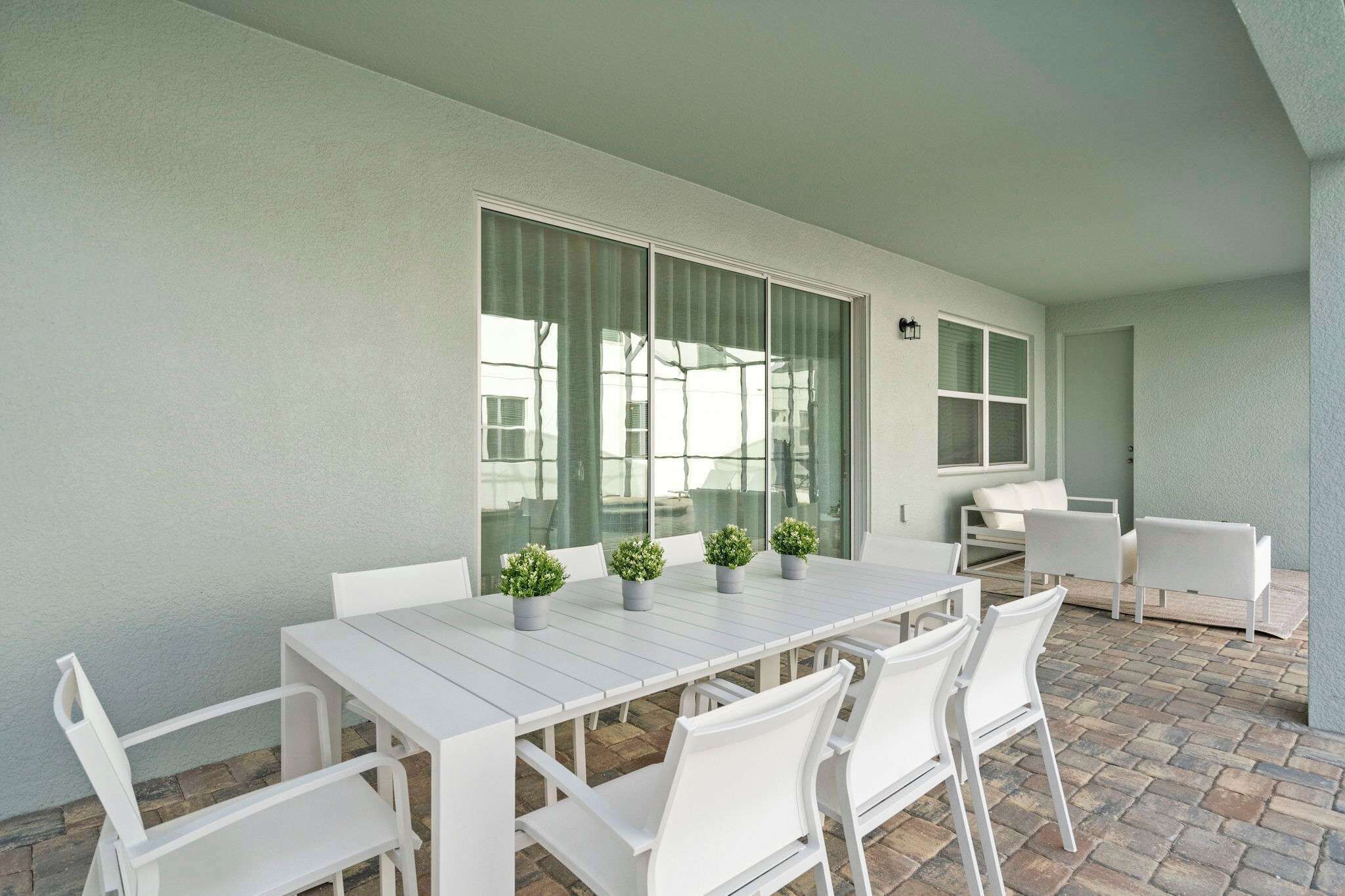 Une terrasse avec une table et des chaises blanches, des plantes en pot et une porte vitrée donnant sur un espace intérieur.