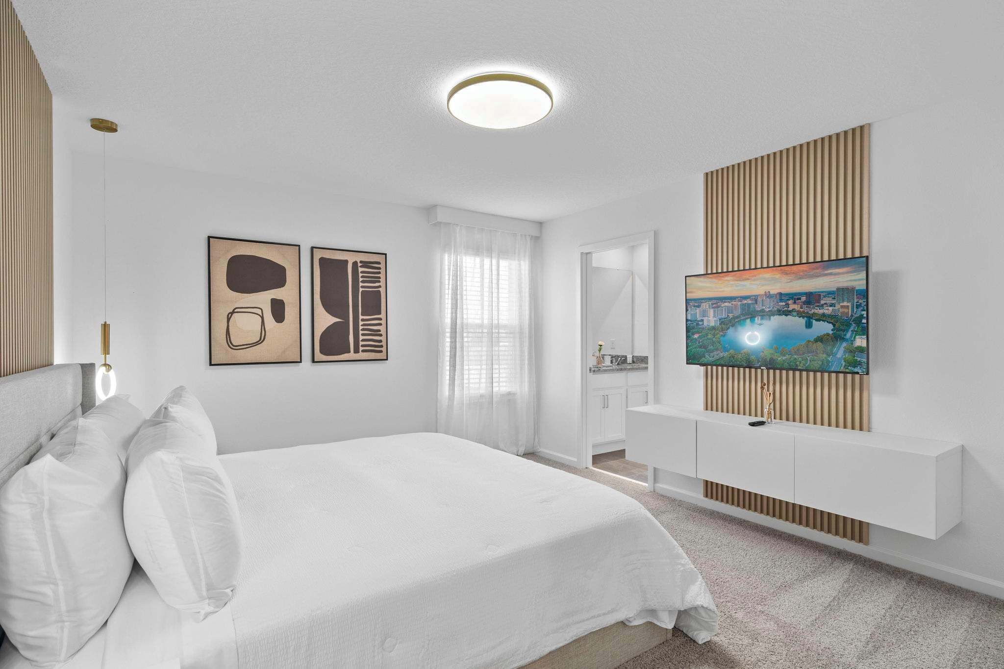 A modern hotel room with a large bed, a TV mounted on a wooden panel, a white wardrobe, and artwork on the wall.