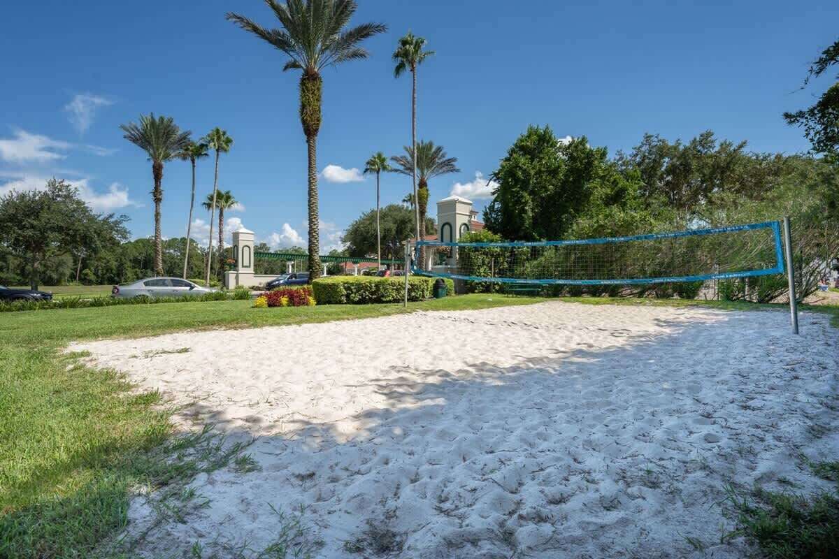 A sandy volleyball court with a blue net, surrounded by palm trees and a building with a green roof in the background.